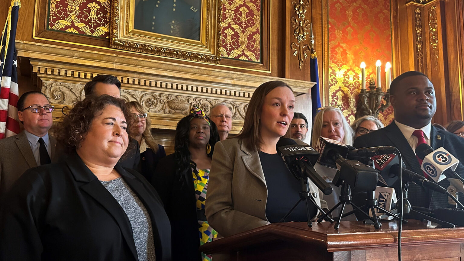 Greta Neubauer stands and speaks into multiple microphones with the flags of different media organizations mounted to the top of a wood podium, with multiple people standing behind her and to her side, in a room with a carved fireplace lintel, a lower portion of a painting, the U.S. and Wisconsin flags, electric wall sconces and toile wallpaper.
