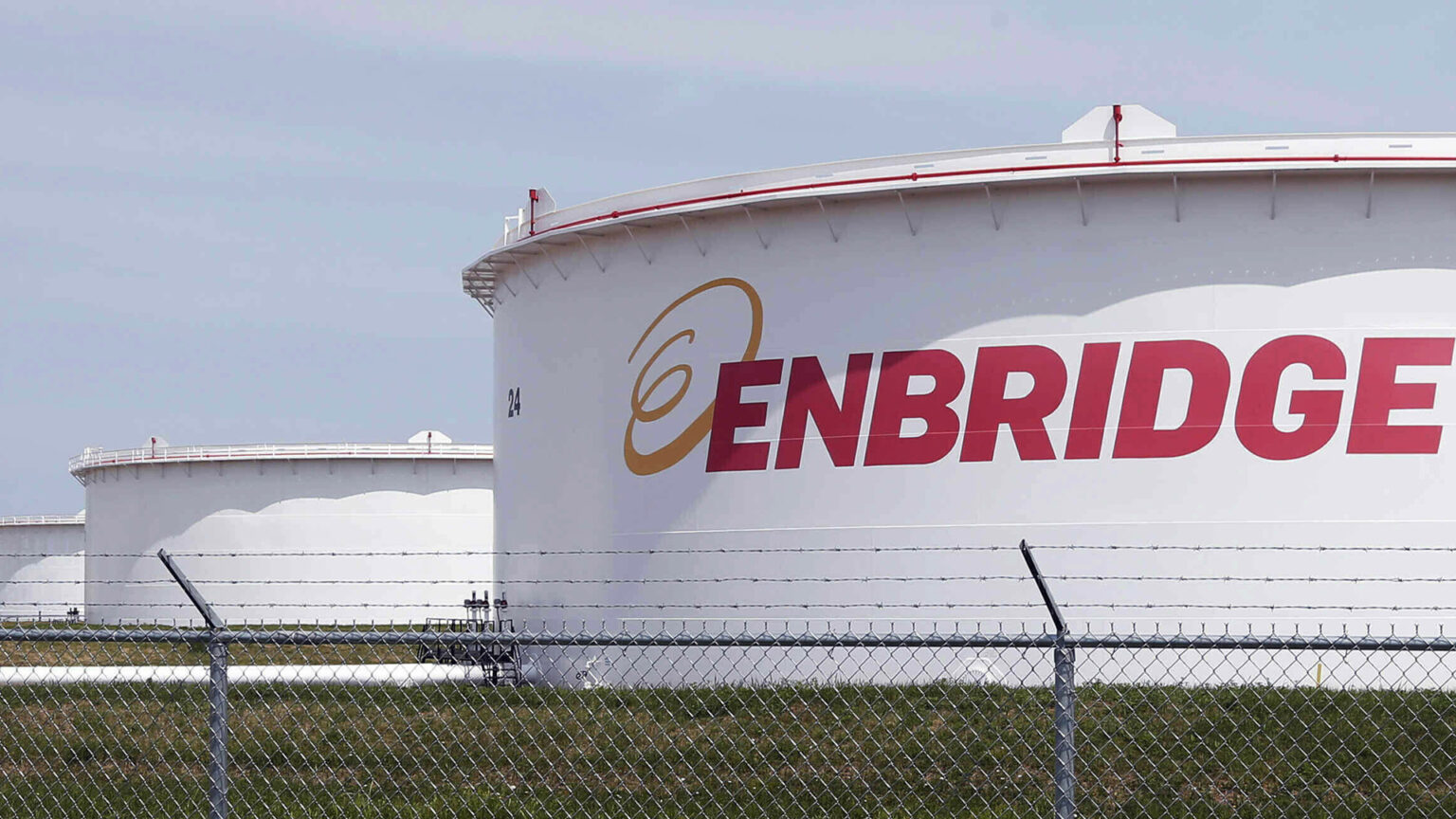 A chain link fence topped by barbed wire stands in front of a gravel road and a row of industrial oil storage tanks with the Enbridge wordmark painted on the side of the one in front.