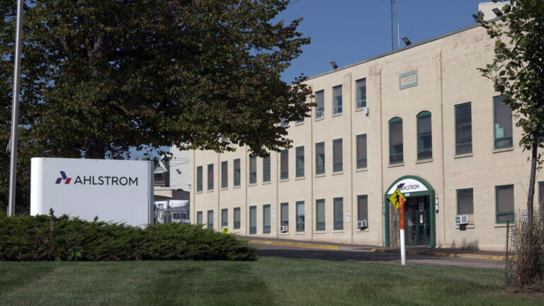 A sign with the Ahlstrom wordmark stands next to a metal flagpole amid landscape plants and in front of a tree with leaves changing colors next to a three-story brick building with rows of windows and an entrance marked by a sign with the word Ahlstrom. A sign with the Ahlstrom wordmark stands next to a metal flagpole amid landscape plants and in front of a tree with leaves changing colors next to a three-story brick building with rows of windows and an entrance marked by a sign with the word Ahlstrom.