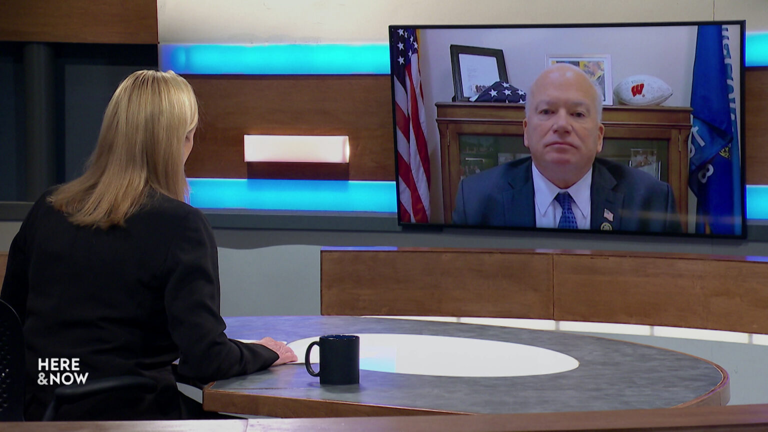 Frederica Freyberg sits at a desk on the Here & Now set and faces a video monitor showing an image of Scott Fitzgerald.