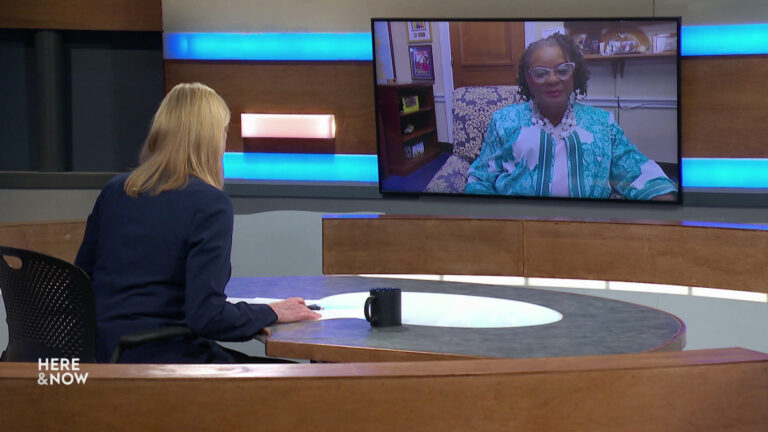 Frederica Freyberg sits at a desk on the Here & Now set and faces a video monitor showing an image of Gwen Moore.