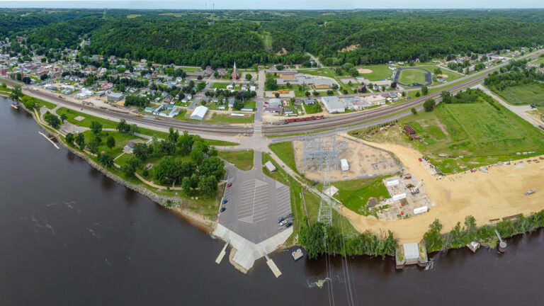 An aerial photo shows a riverfront parking lot with two docks in between a wooded area and open fields with an electrical substation connected to power lines spanning water, with two railroad lines and a landscape of roads and houses at the foot of wooded bluffs and more fields and wooded areas leading to the horizon.