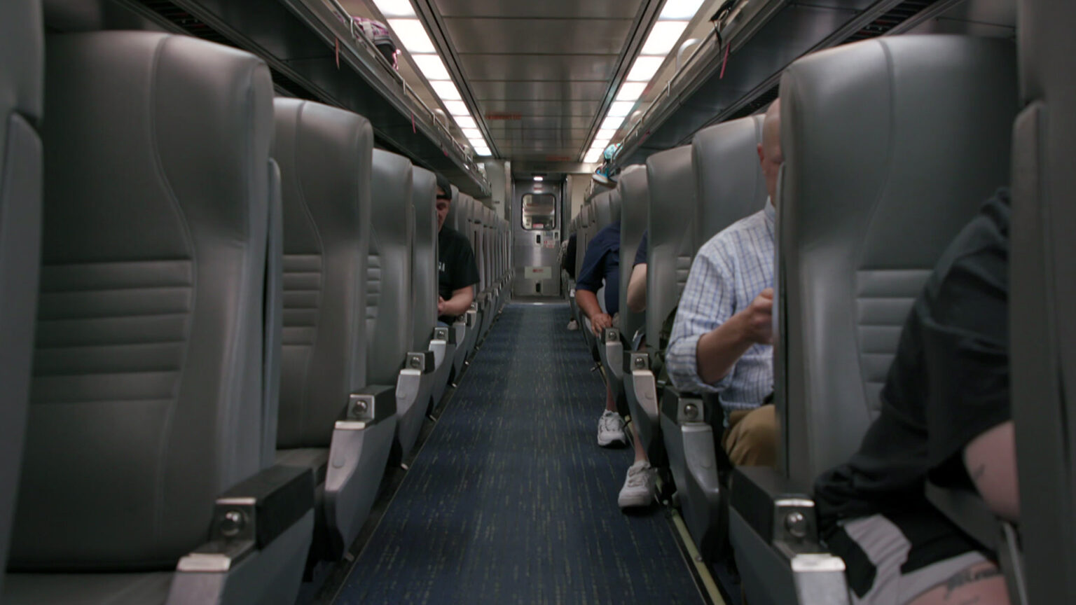 Passengers sit in multiple seats on either side of an aisle in a train car with an industrial carpet and two lines of overhead lighting.