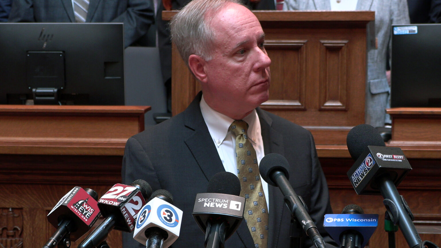 Robin Vos stands and listens while standing in front of multiple microphones with the flags of different media organizations, with a podium, multiple computer monitors and people standing on a legislative dais in the background. Robin Vos stands and listens while standing in front of multiple microphones with the flags of different media organizations, with a podium, multiple computer monitors and people standing on a legislative dais in the background.