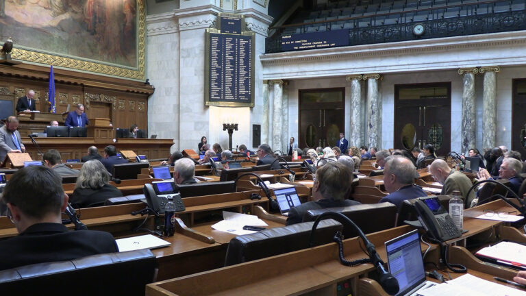 A lawmaker stands and speaks into a microphone mounted to a wood podium at the center of a legislative dais, with multiple people seated in high-backed wood and leather chairs beside and behind him on the dais, faced by more people seated in multiple rows of wood desks, in a room with a large painting and taxidermy eagle on the rear wall, an electronic vote register on an adjacent wall, and marble pillars supporting a second-level seating gallery.