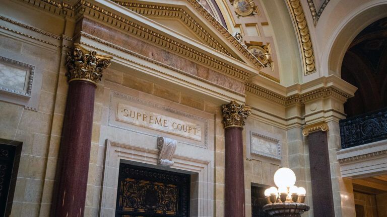 Marble pillars with Composite order capitals frame a doorway in a marble masonry wall with a carved sign reading Supreme Court above a metal filigree door, with additional doors on either side, a pediment above, and an illuminated brass pillar electric light fixture in the foreground. Marble pillars with Composite order capitals frame a doorway in a marble masonry wall with a carved sign reading Supreme Court above a metal filigree door, with additional doors on either side, a pediment above, and an illuminated brass pillar electric light fixture in the foreground.