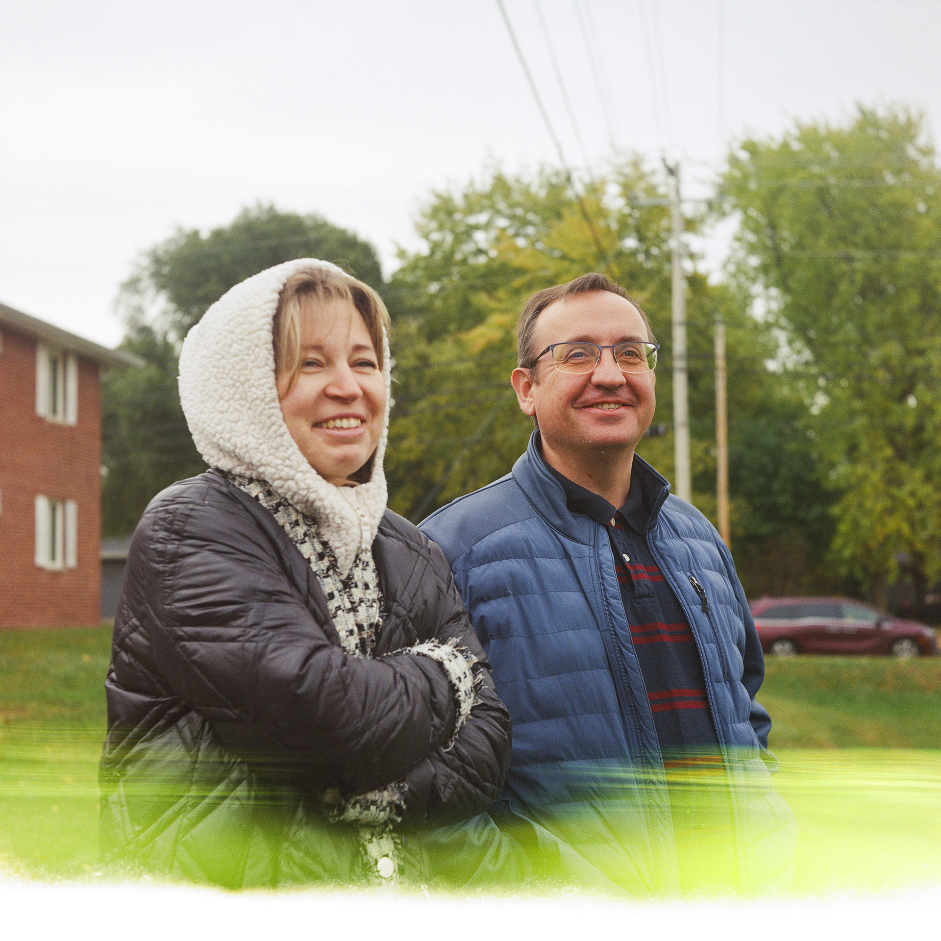 Anna Mykhailova and Sasha Druzhyna smile while standing outside in front of a building, a parked vehicle, trees, and utility poles and lines, with a horizontal light distortion at the bottom of the image.