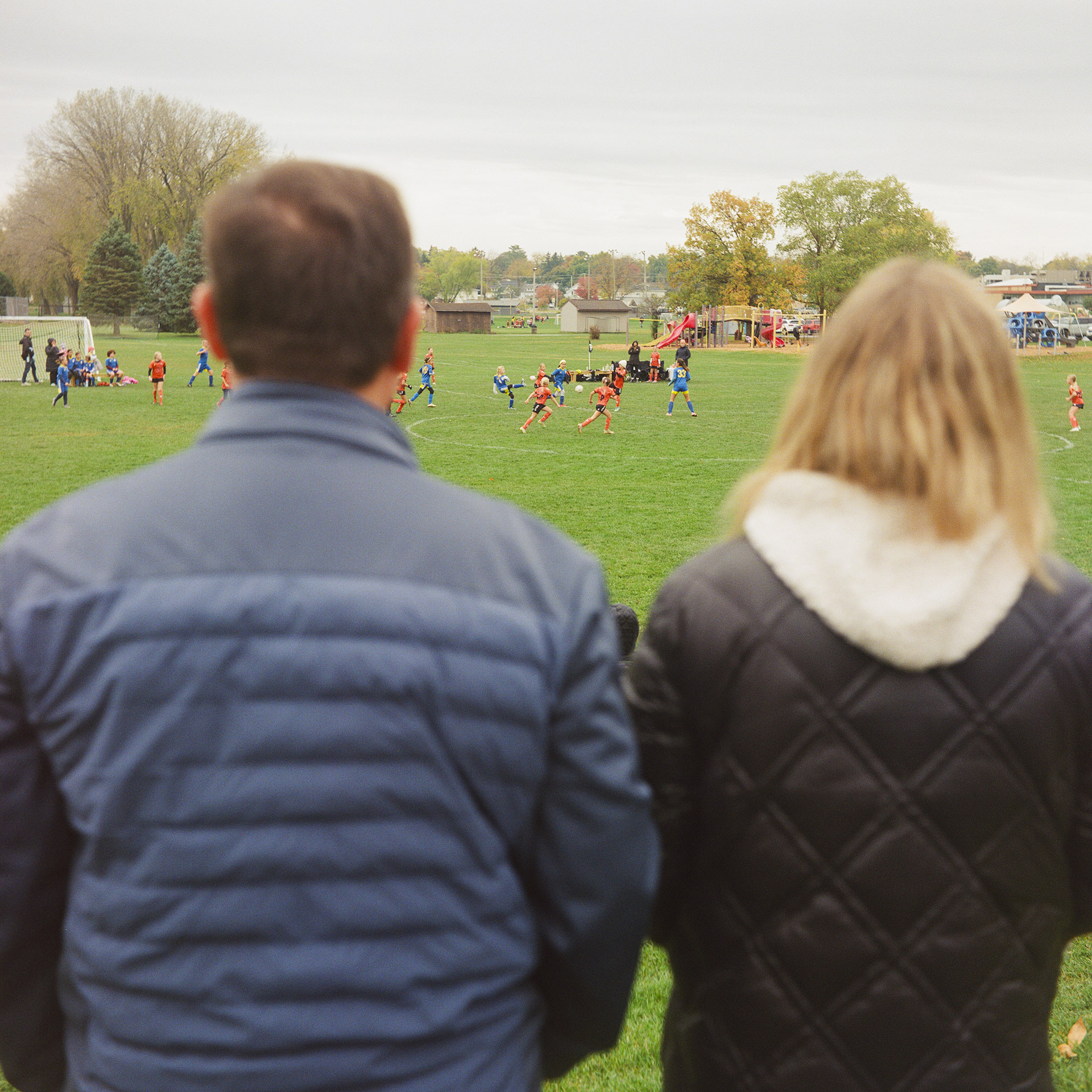 An out-of-focus Sasha Druzhyna and Anna Mykhailova face away from the camera and toward a youth soccer game on a field, with playground equipment, trees and buildings along the horizon.