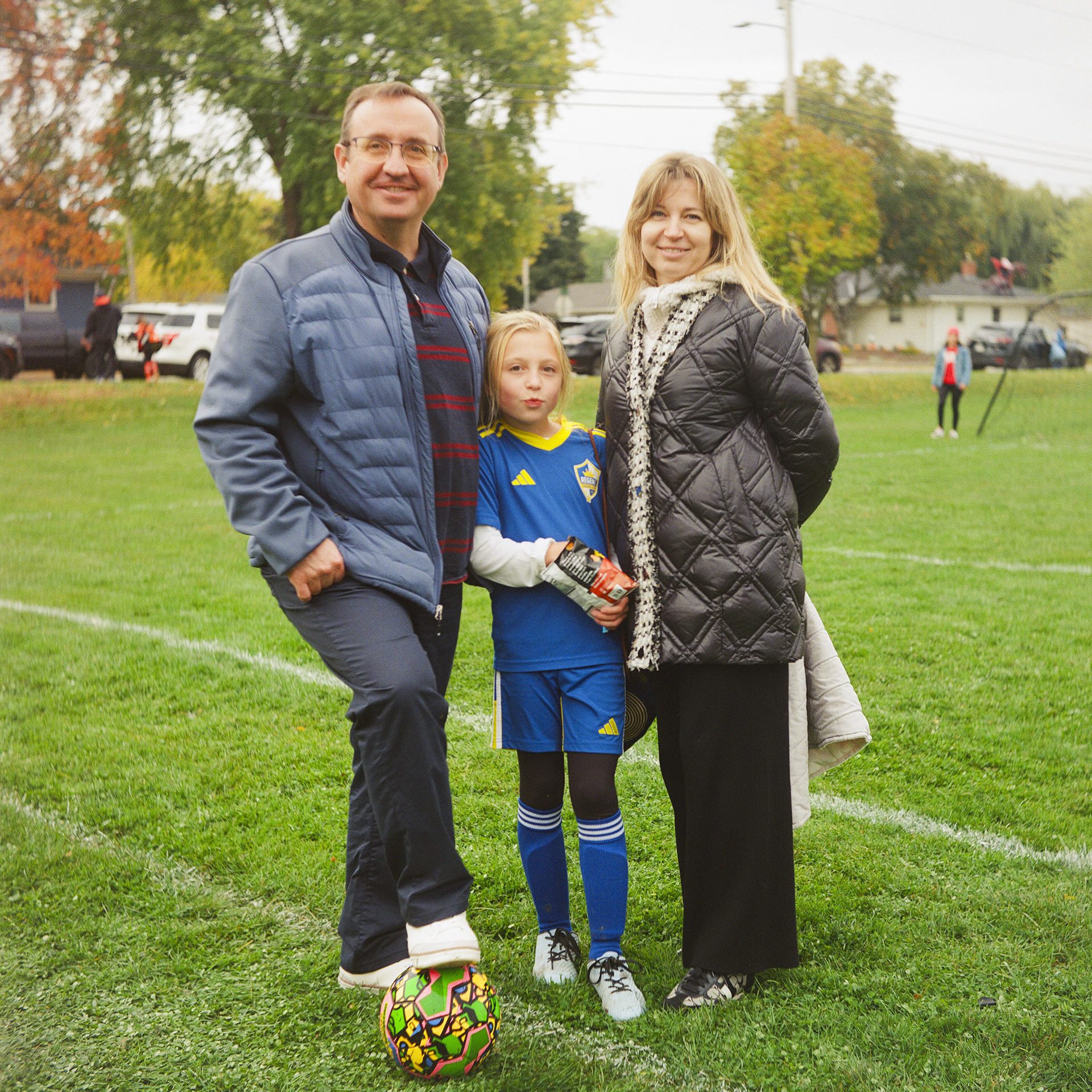 Sasha Druzhyna, Varya Mykhailova and Anna Mykhailova pose for a portrait outside on a soccer field, with Sasha's right foot standing on a soccer ball and with out-of-focus trees, parked vehicles and houses in the background.