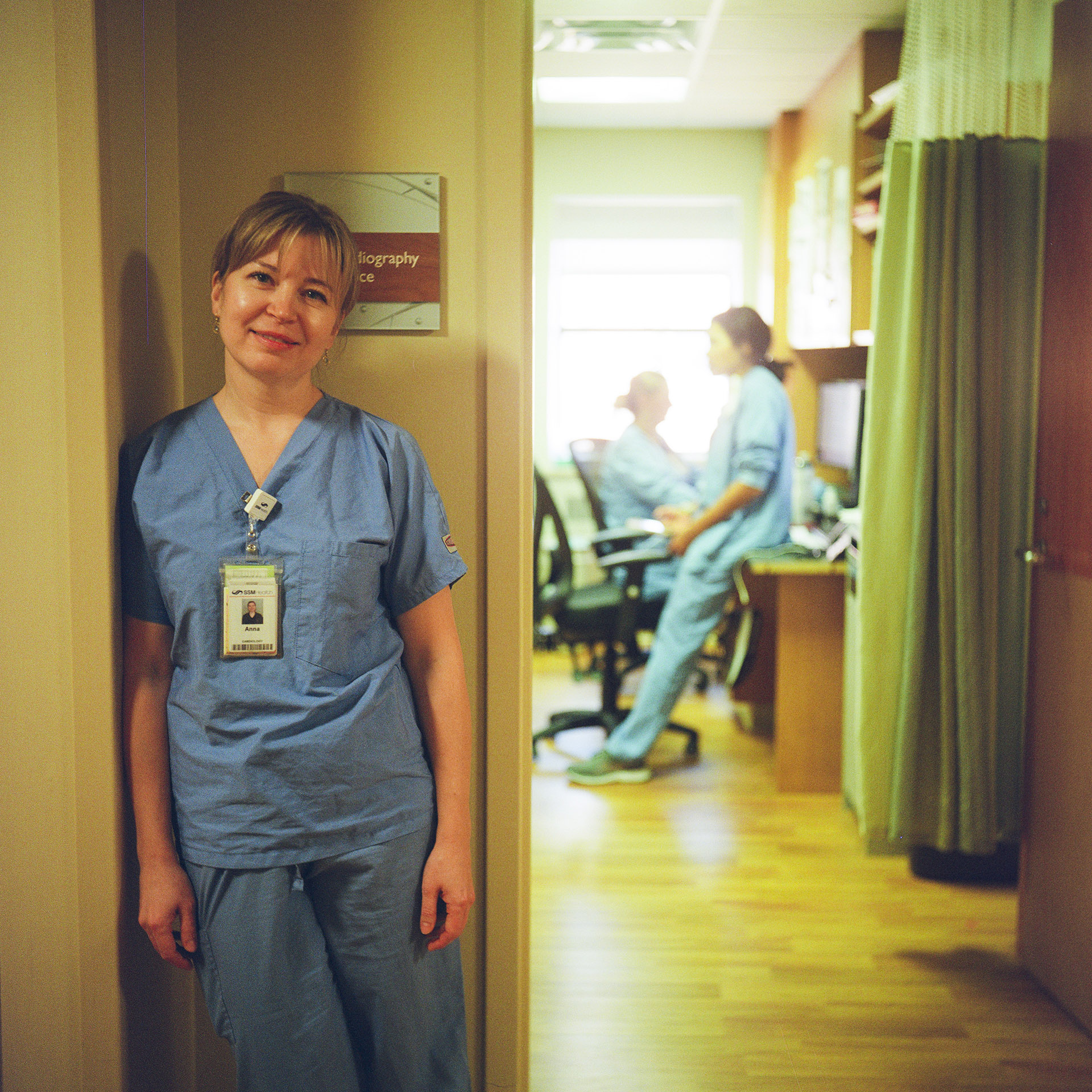 Anna Mykhailova poses for a portrait while standing in a corner next to an open door to a room with a privacy curtain and two out-of-focus people sitting in a rolling chair and on the surface of a desk in front of a sunlight-illuminated window.
