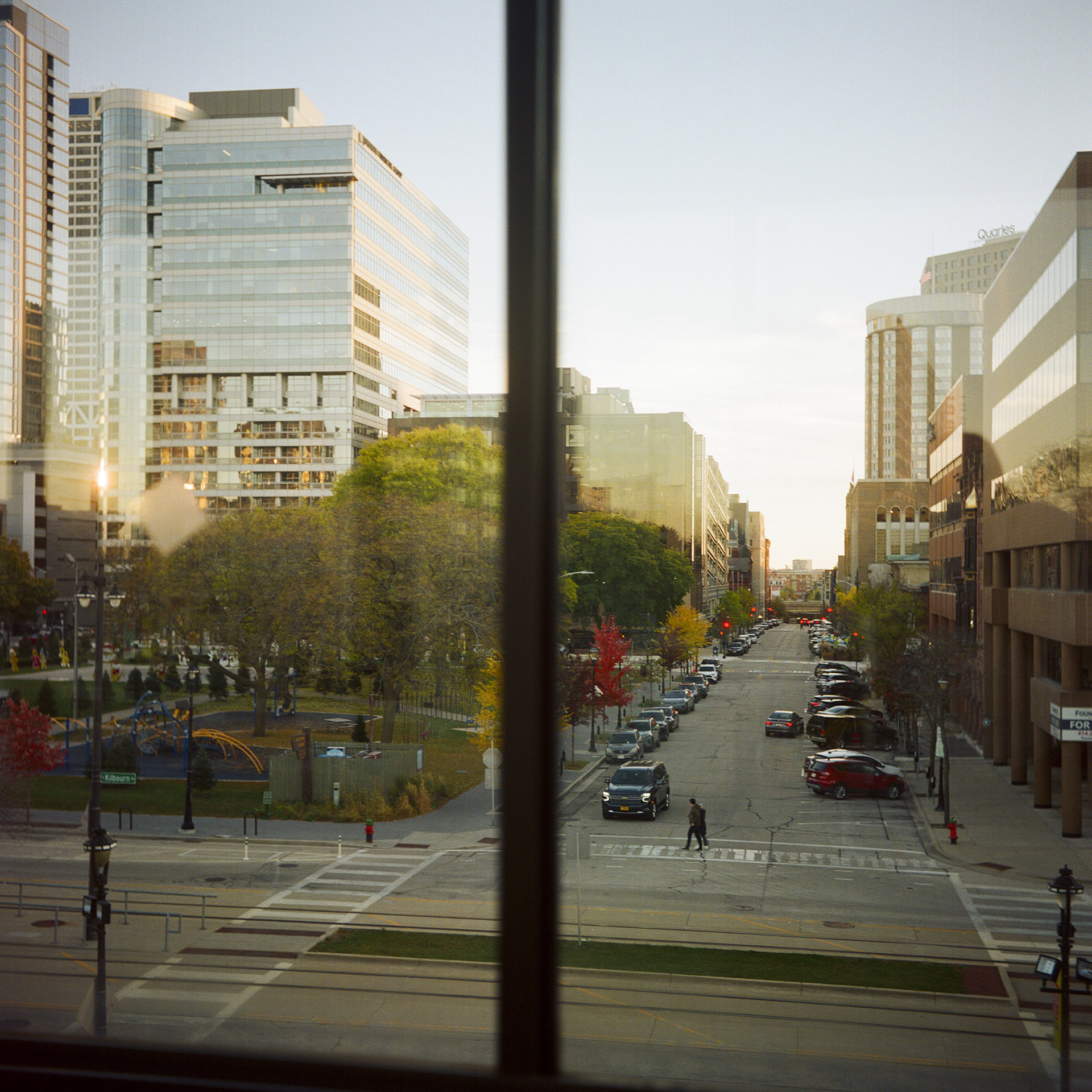 A pedestrian crosses a street in front of a vehicle at a T-intersection in an urban landscape with buildings of varying heights and designs, vehicles parked on the side of a street and trees with different sizes, as seen through the window of another building.