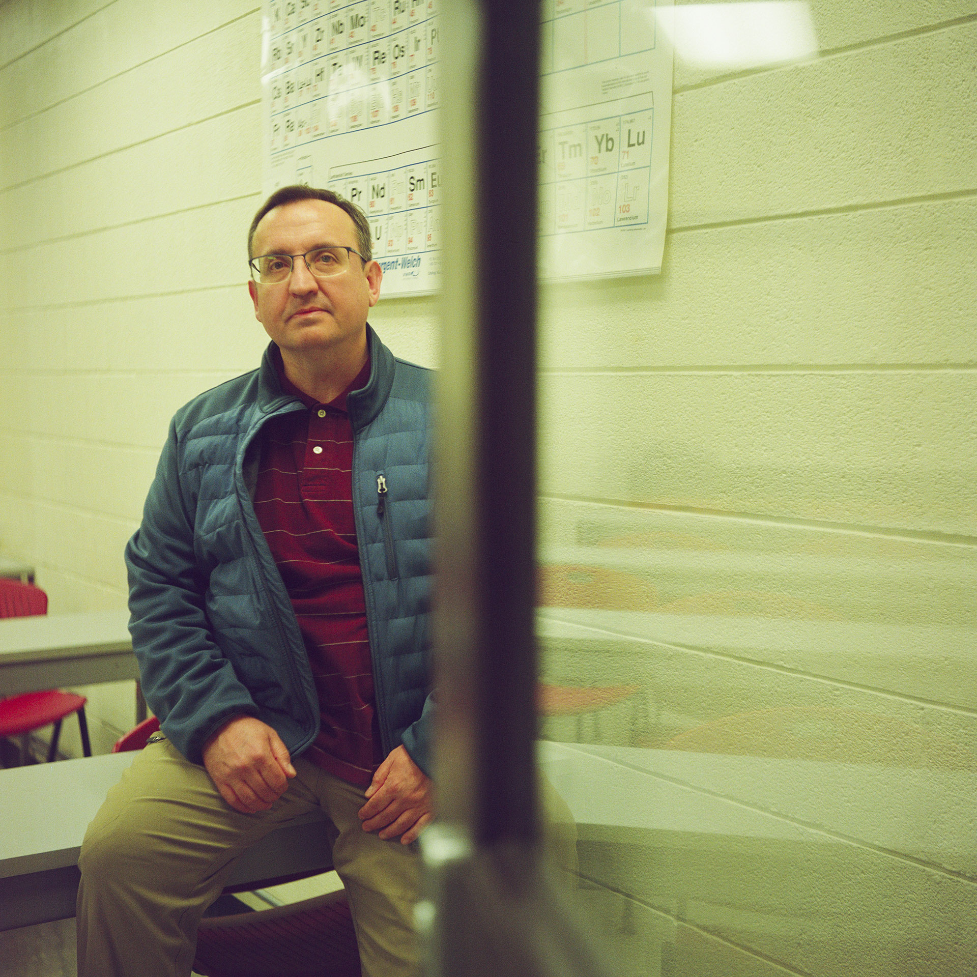 Sasha Druzhyna poses for a portrait while seated at the edge of a table in front of a periodic table of elements displayed on a painted concrete-block wall, with an out-of-focus glass-and-metal door on the right side of the foreground.