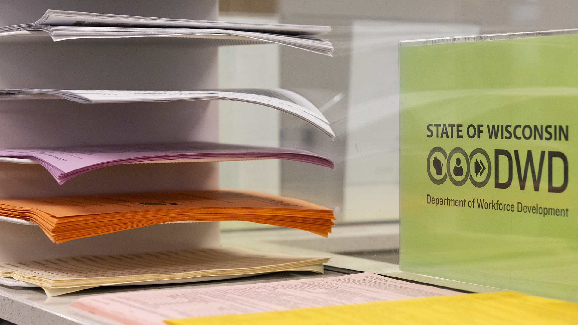 A printed paper sign with the text "State of Wisconsin" and "Department of Workforce Development above and below a DWD wordmark is encased in a transparent display stand next to a flat surface with multiple stacks of paper and a paper filing stand with stacks of paper in each slot.