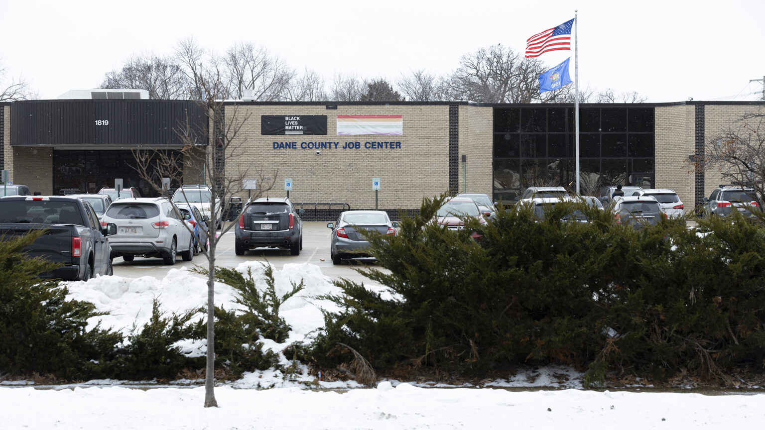 The U.S. and Wisconsin flags fly from the top of a flagpole in front of a single-story building with brick and glass walls and a letter sign reading Dane County Job Center beneath Black Lives Matter and the rainbow Pride flag banners, with multiple vehicles parked in a parking lot in front of the building, and with snowbanks, leafless trees and evergreen landscape plants in the foreground, and the tops of leafless trees behind the building in the background.