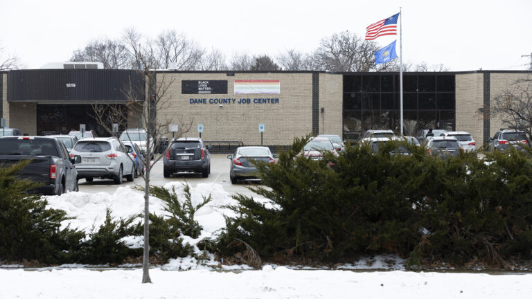 The U.S. and Wisconsin flags fly from the top of a flagpole in front of a single-story building with brick and glass walls and a letter sign reading Dane County Job Center beneath Black Lives Matter and the rainbow Pride flag banners, with multiple vehicles parked in a parking lot in front of the building, and with snowbanks, leafless trees and evergreen landscape plants in the foreground, and the tops of leafless trees behind the building in the background. The U.S. and Wisconsin flags fly from the top of a flagpole in front of a single-story building with brick and glass walls and a letter sign reading Dane County Job Center beneath Black Lives Matter and the rainbow Pride flag banners, with multiple vehicles parked in a parking lot in front of the building, and with snowbanks, leafless trees and evergreen landscape plants in the foreground, and the tops of leafless trees behind the building in the background.