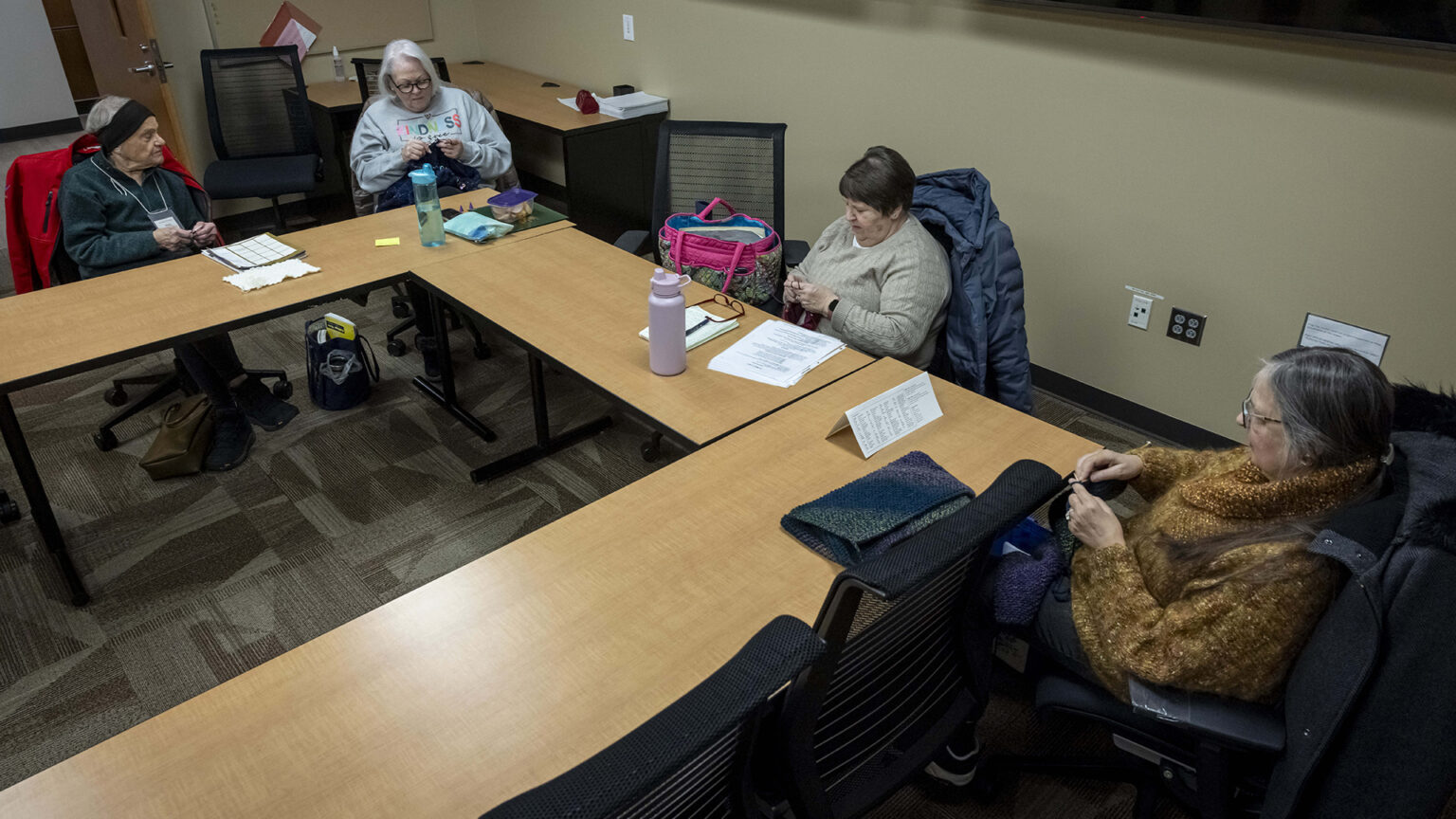 Anita Kirschling, Theresa Reiter, Judy Rogers and Linda Chapman knit while sitting on rolling office chairs around three sides of tables arranged in a square, with other chairs facing the tables and in the background in a room with an industrial carpet and an open wood door.