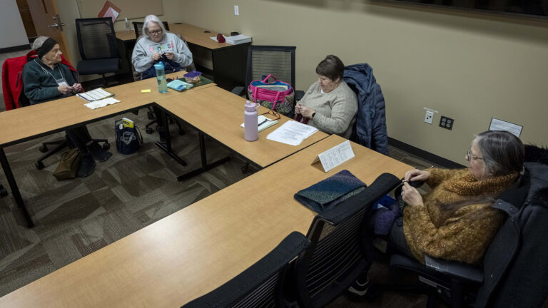 Anita Kirschling, Theresa Reiter, Judy Rogers and Linda Chapman knit while sitting on rolling office chairs around three sides of tables arranged in a square, with other chairs facing the tables and in the background in a room with an industrial carpet and an open wood door.