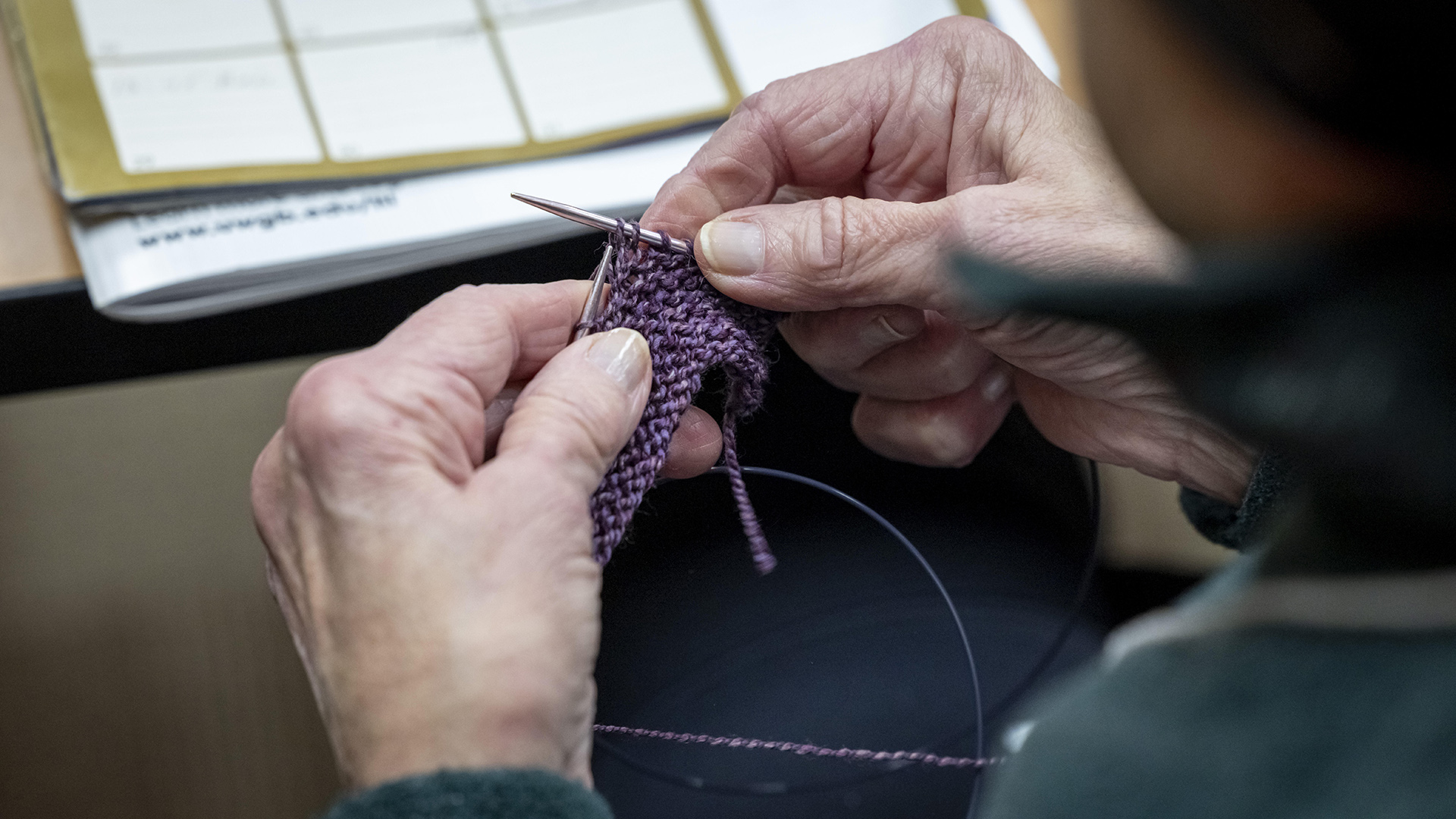 Anita Kirschling's hands holds knitting needles while working on a fiber art project above out of-focus books on the surface of a table.