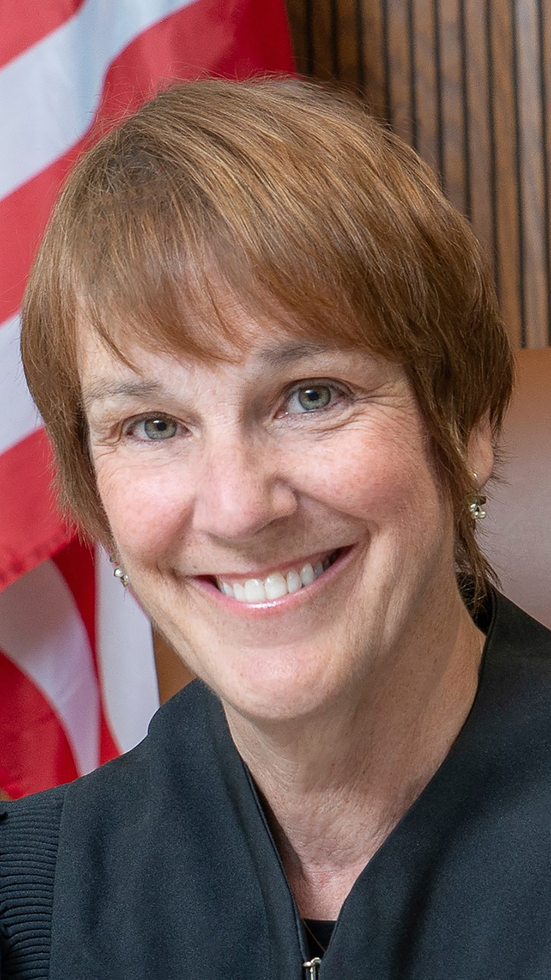 Lisa Neubauer poses for a portrait while wearing judicial robes and with the stripes of a U.S. flag in the background.
