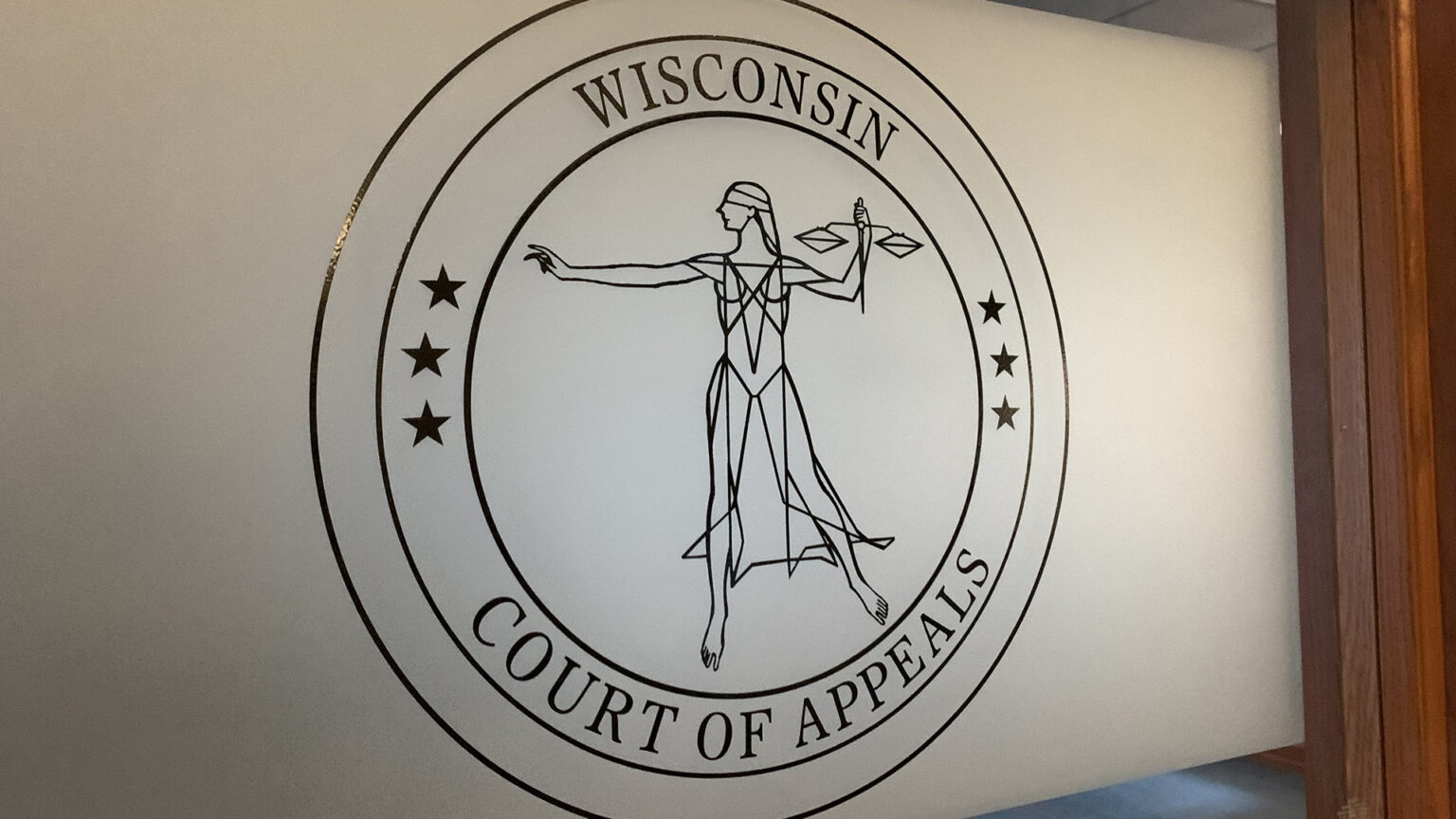 A circular seal showing a stylized Lady Justice surrounded by two sets of three five-pointed stars and the words Wisconsin Court of Appeals is displayed on a frosted window in a wood frame. A circular seal showing a stylized Lady Justice surrounded by two sets of three five-pointed stars and the words Wisconsin Court of Appeals is displayed on a frosted window in a wood frame.