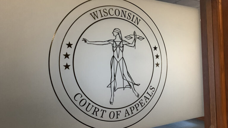 A circular seal showing a stylized Lady Justice surrounded by two sets of three five-pointed stars and the words Wisconsin Court of Appeals is displayed on a frosted window in a wood frame.