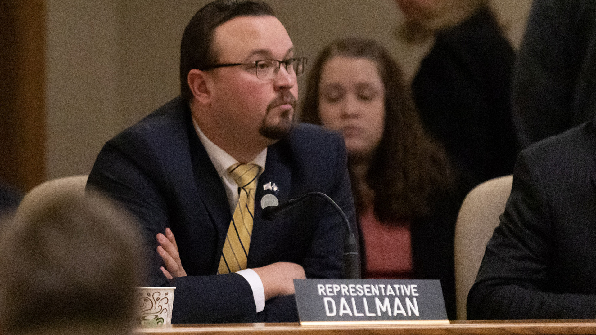 Alex Dallman sits with his arms crossed in front of him at a legislative dais in front of a mounted microphone, paper cup and a sign reading "Representative Dallman," with another person seated to his side and out-of-focus people in the foreground and background.