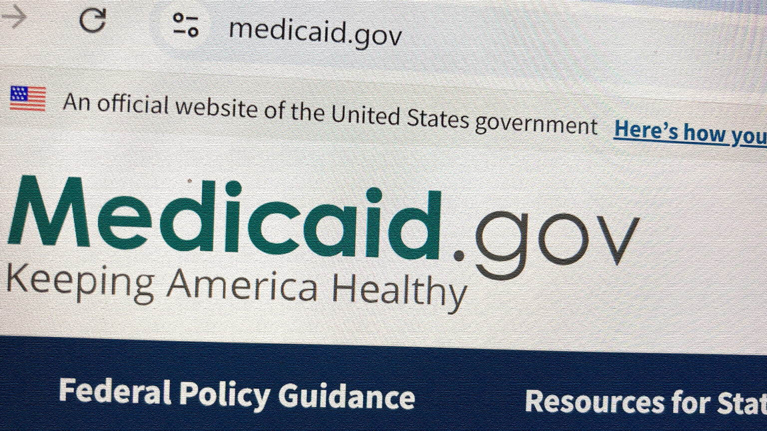 A photo shows a website displayed on a Google Chrome browser with a URL bar reading medicaid.gov above lines of text reading An official website of the United States government, Medicaid.gov, Keeping America Healthy, Federal Policy Guidance and other partial phrases.