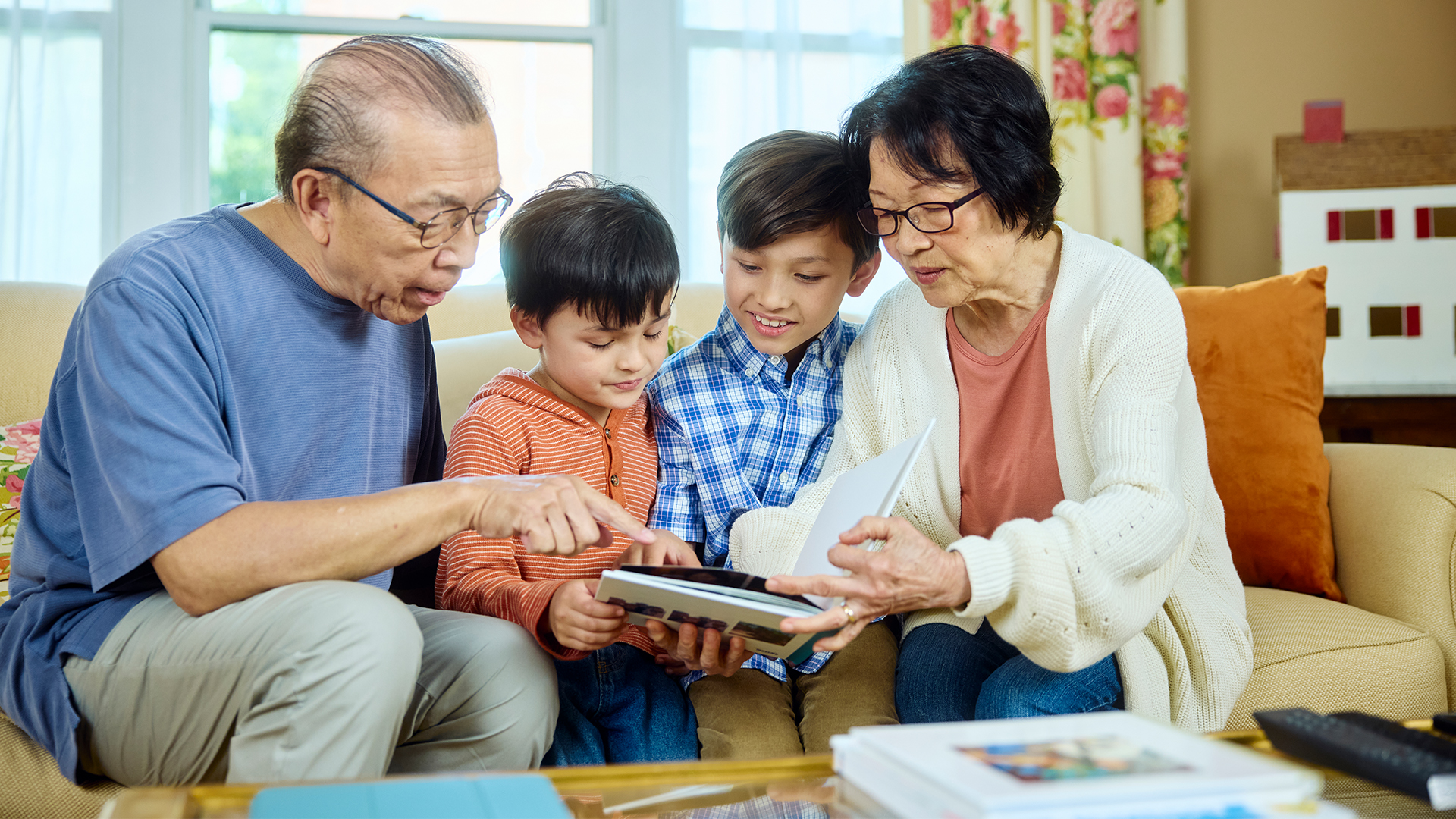 Grandparents and two children sit on a cozy sofa, sharing a book. They are smiling, creating a warm, familial atmosphere in a bright, inviting living room.