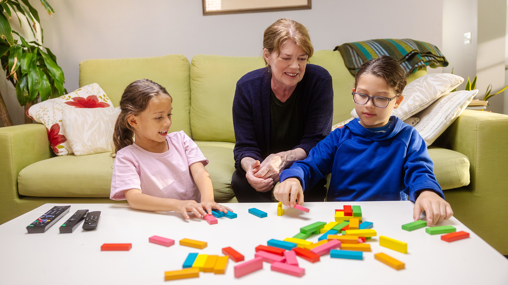 A woman and two children play with colorful blocks on a white table in a cozy living room, creating a joyful and focused atmosphere.