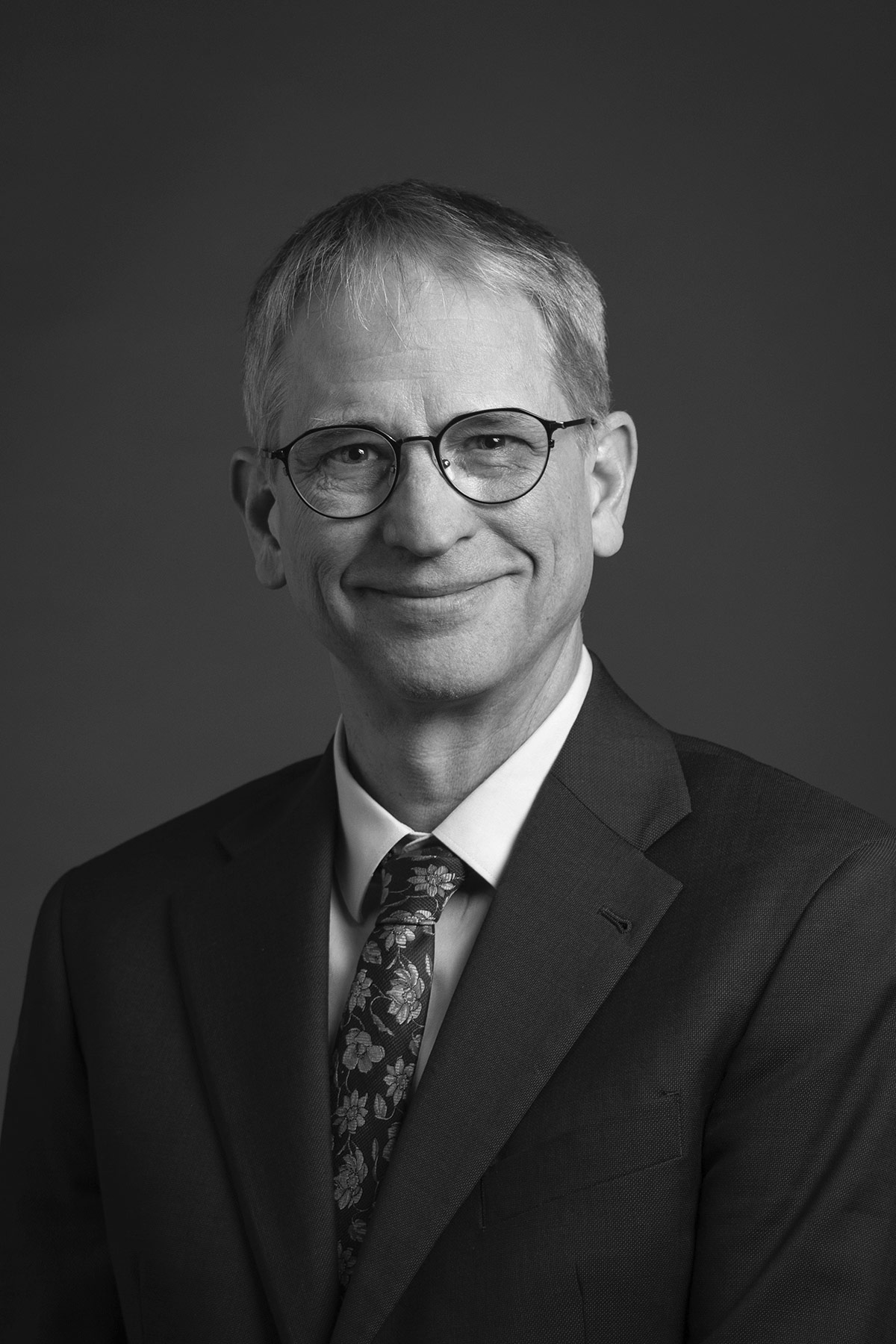 Black-and-white headshot of an older man wearing glasses, suit, and patterned tie, smiling at the camera.