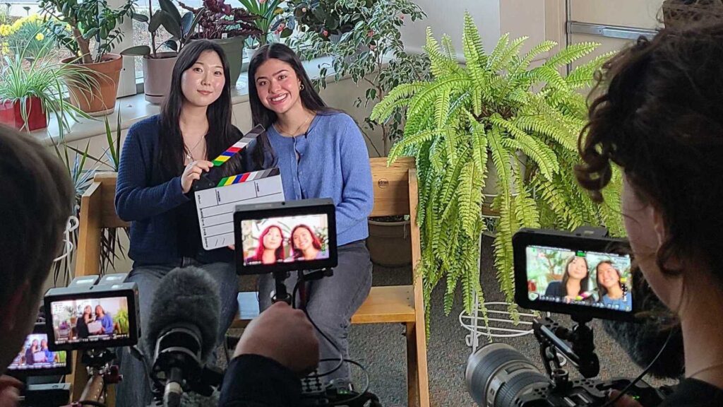 Two young women sit together while a film crew sets up an interview.