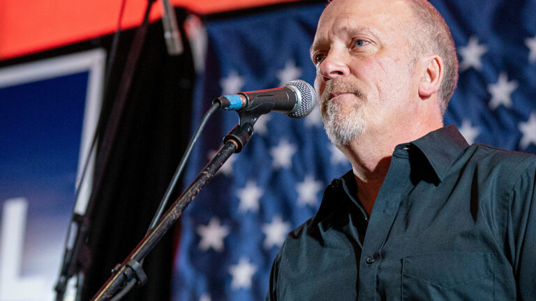 Brad Schimel speaks into a microphone mounted on a stand while standing in front of an out-of-focus U.S. flag.