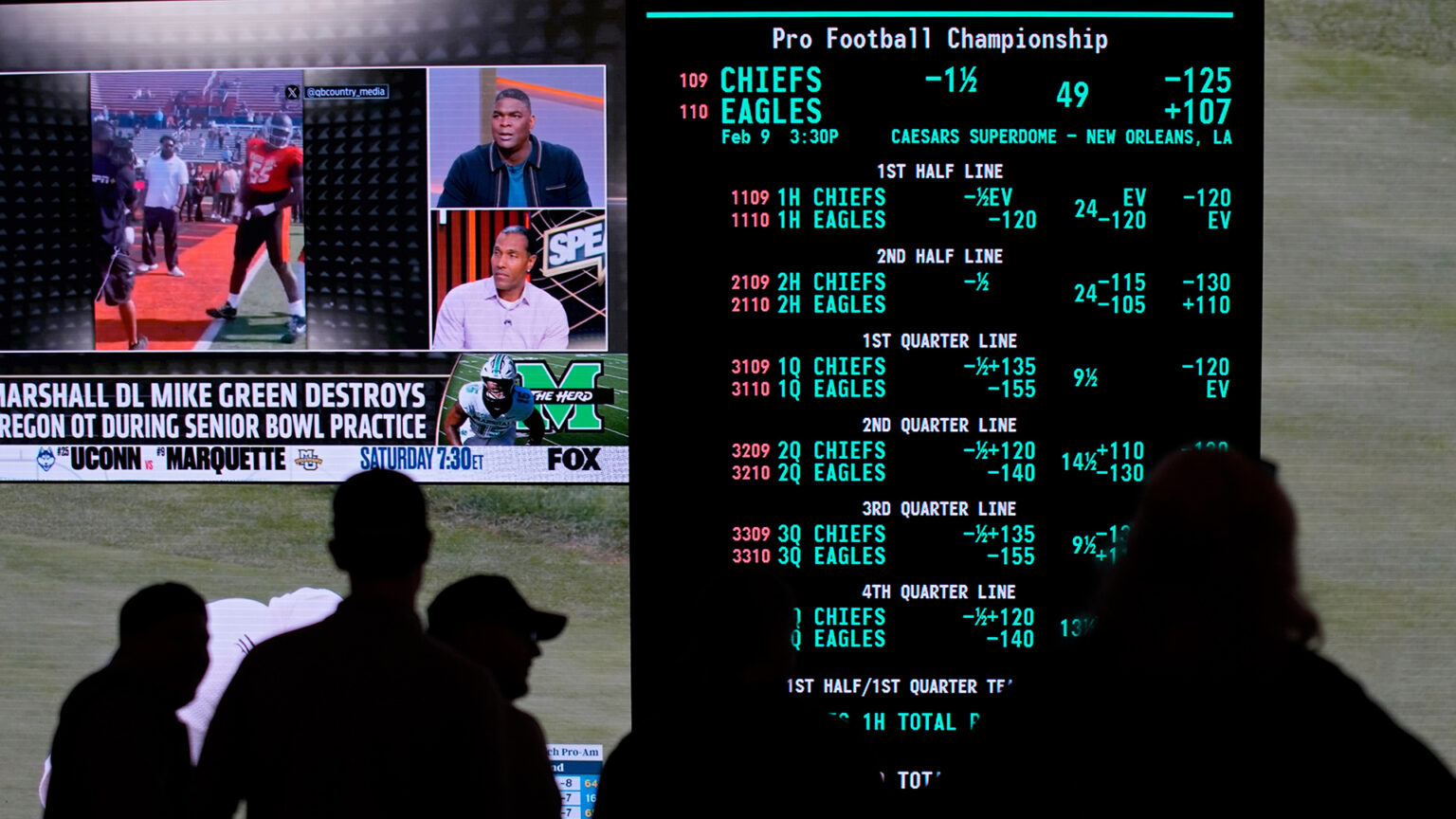 A screen shows multiple lines for the Pro Football Championship between the Chiefs and Eagles, with an adjacent screen showing a sports broadcast on the FOX network, with the silhouettes of standing people in the foreground.