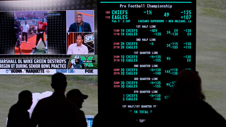 A screen shows multiple lines for the Pro Football Championship between the Chiefs and Eagles, with an adjacent screen showing a sports broadcast on the FOX network, with the silhouettes of standing people in the foreground.