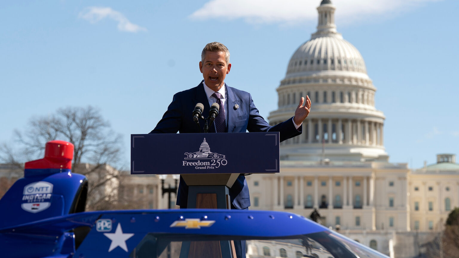 Sean Duffy stands and gestures with his left hand while speaking into two microphones mounted to the top of a podium with a placard of an illustration showing an IndyCar in front of a dome with the words Freedom 250 Grand Prix mounted to its front, with an out-of-focus top of an IndyCar with a Chevrolet logo in the foreground, and an out-of-focus neoclassical masonry building with two wings flanking a central dome. Sean Duffy stands and gestures with his left hand while speaking into two microphones mounted to the top of a podium with a placard of an illustration showing an IndyCar in front of a dome with the words Freedom 250 Grand Prix mounted to its front, with an out-of-focus top of an IndyCar with a Chevrolet logo in the foreground, and an out-of-focus neoclassical masonry building with two wings flanking a central dome.