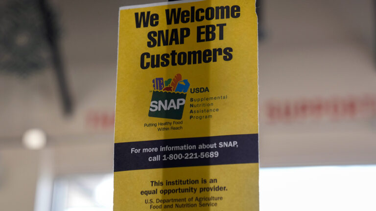 A printed paper sign with text reading We Welcome SNAP EBT Customers, the SNAP logo, a line pointing to a phone number and other text is displayed inside a grocery store.