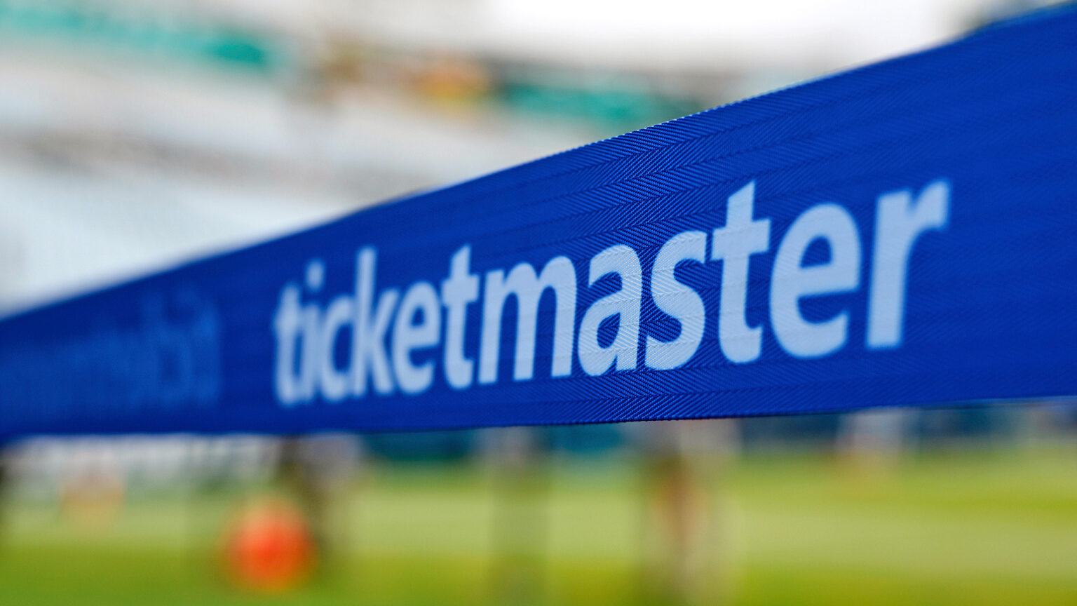 The Ticketmaster wordmark is printed on the surface of a retractable belt barrier, with an out-of-focus grass turf and grandstand in the background.