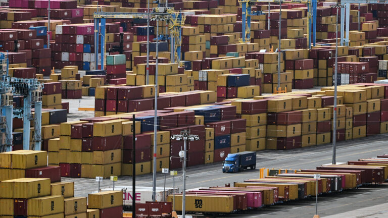 A semi-truck drives on a lane between a row of semi-trailers and numerous stacks of shipping containers positioned around multiple cranes.