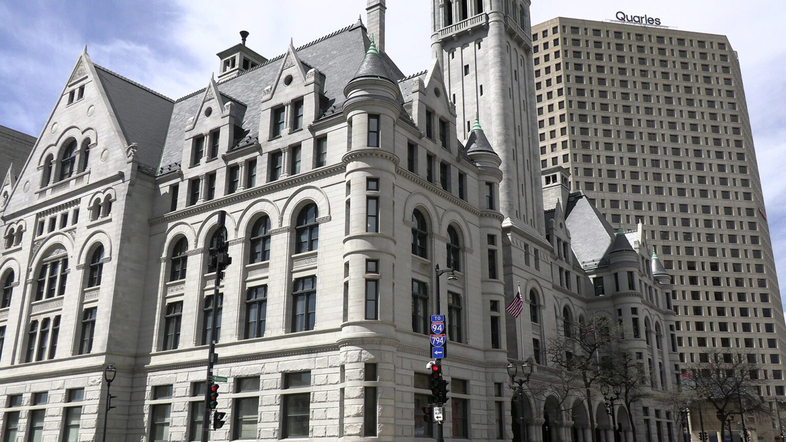 Sunlight illuminates and casts shadows on a multi-story Richardsonian Romanesque-style masonry building with multiple turrets, gables and a central tower, with two U.S. flags flying on flagpoles next to leafless trees at its central entrance and with other buildings behind it.