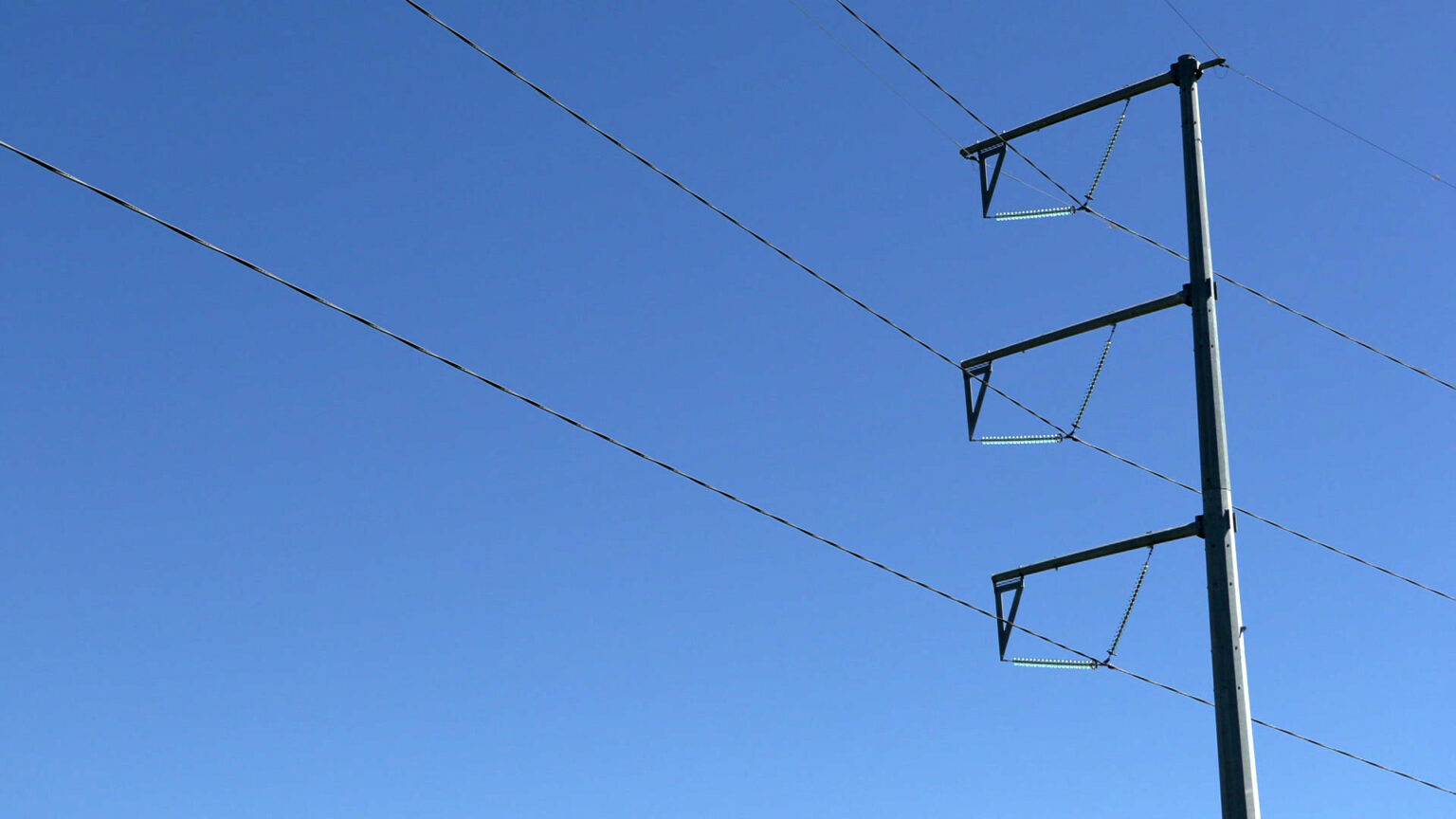 Sunlight reflects off one side of a metal utility pole with three tiers and electricity transmission lines running through each, with a clear sky in the background.
