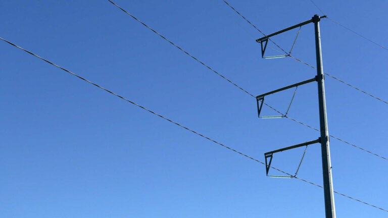 Sunlight reflects off one side of a metal utility pole with three tiers and electricity transmission lines running through each, with a clear sky in the background. Sunlight reflects off one side of a metal utility pole with three tiers and electricity transmission lines running through each, with a clear sky in the background.