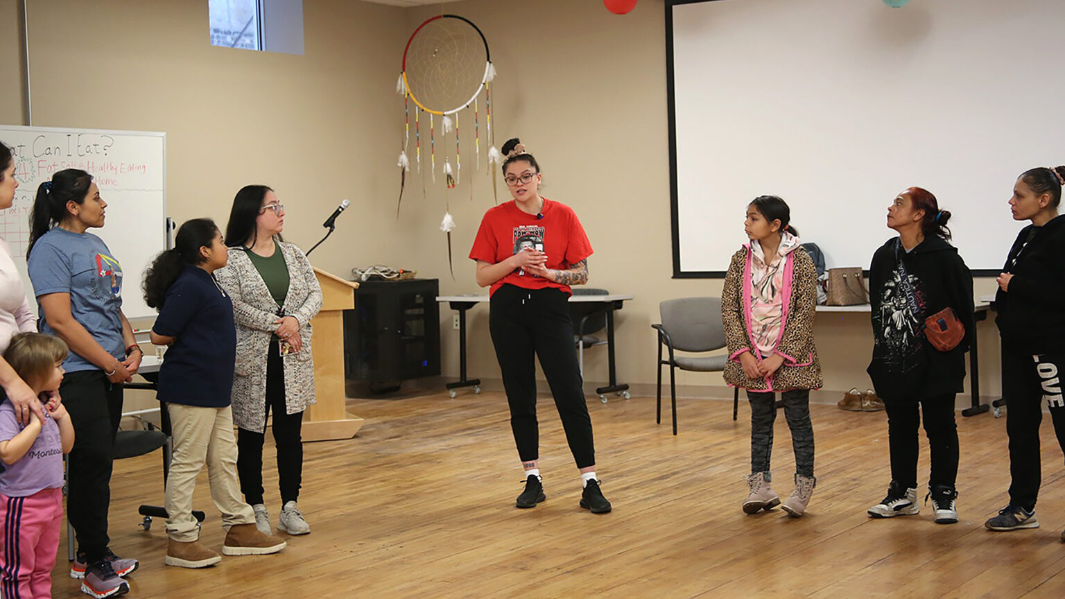 Emma Carufel stands on a wood floor, with lines of people standing on either side of her, in a room with chairs, tables, a podium with a microphone and a whiteboard in the background along with a larger projector screen on a wall and a ceiling-mounted large dream catcher displayed in a corner.