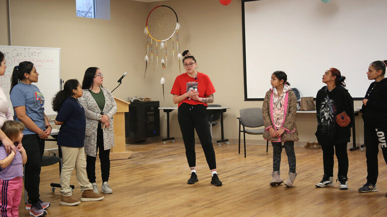 Emma Carufel stands on a wood floor, with lines of people standing on either side of her, in a room with chairs, tables, a podium with a microphone and a whiteboard in the background along with a larger projector screen on a wall and a ceiling-mounted large dream catcher displayed in a corner.