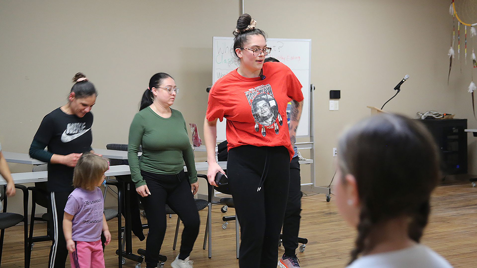 Emma Carufel dances with other people dancing behind her and the back of the head of an out-of-focus person facing her in the foreground, in a room with a wood floor, stackable chairs and tables, a whiteboard, and a ceiling-mounted large dream catcher.