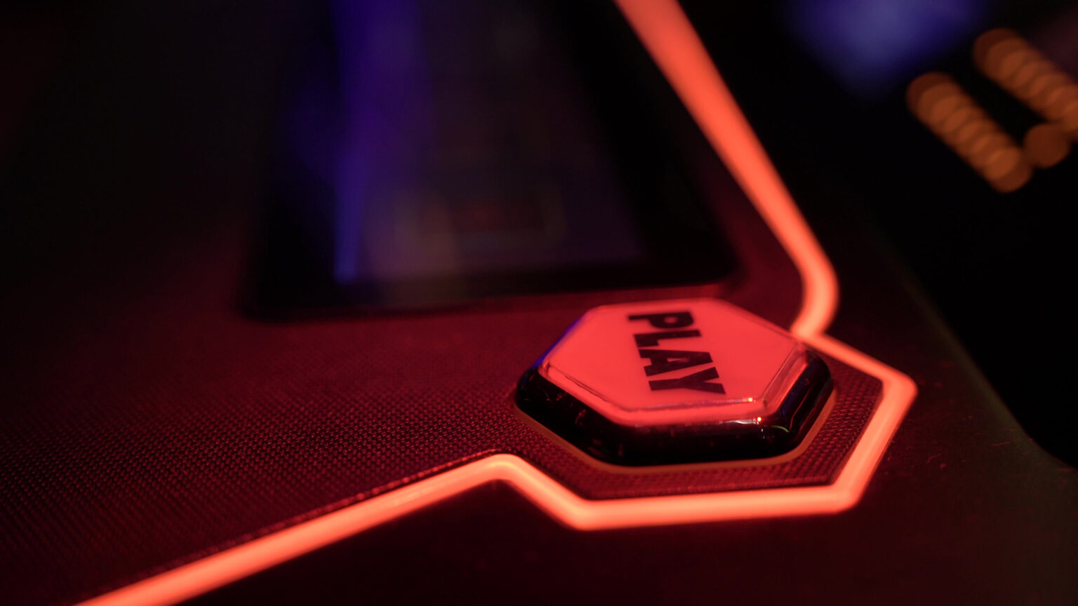 Light illuminates a plastic hexagonal button with the text PLAY on its surface that is surrounded by illuminated piping on the interface of a slot machine.