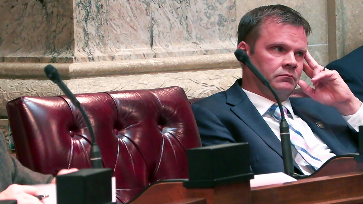 Devin LeMahieu holds his left hand's index and middle fingers to the left side of his face while seating at a desk, with a high-backed leather chair to his side and other people seated on either side in a room with marble masonry.