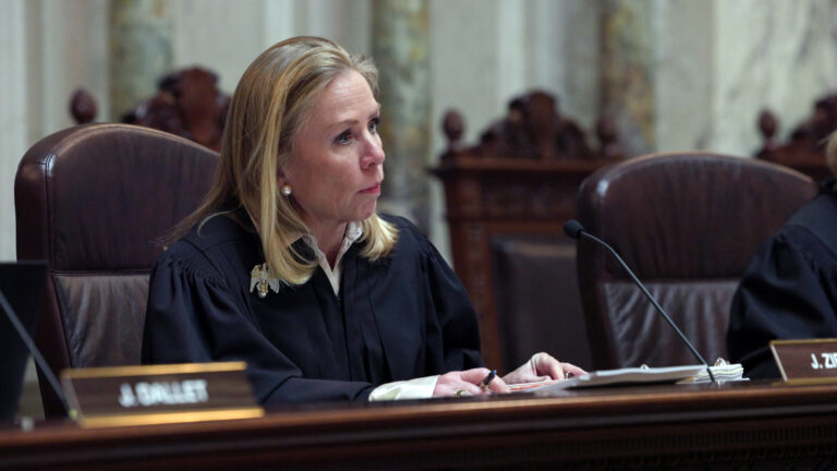 Annette Ziegler listens while sitting in a high-backed leather chair and facing a microphone mounted to a judicial dais with an open binder filled with papers on its surface, with other high-backed wood chairs in the background.