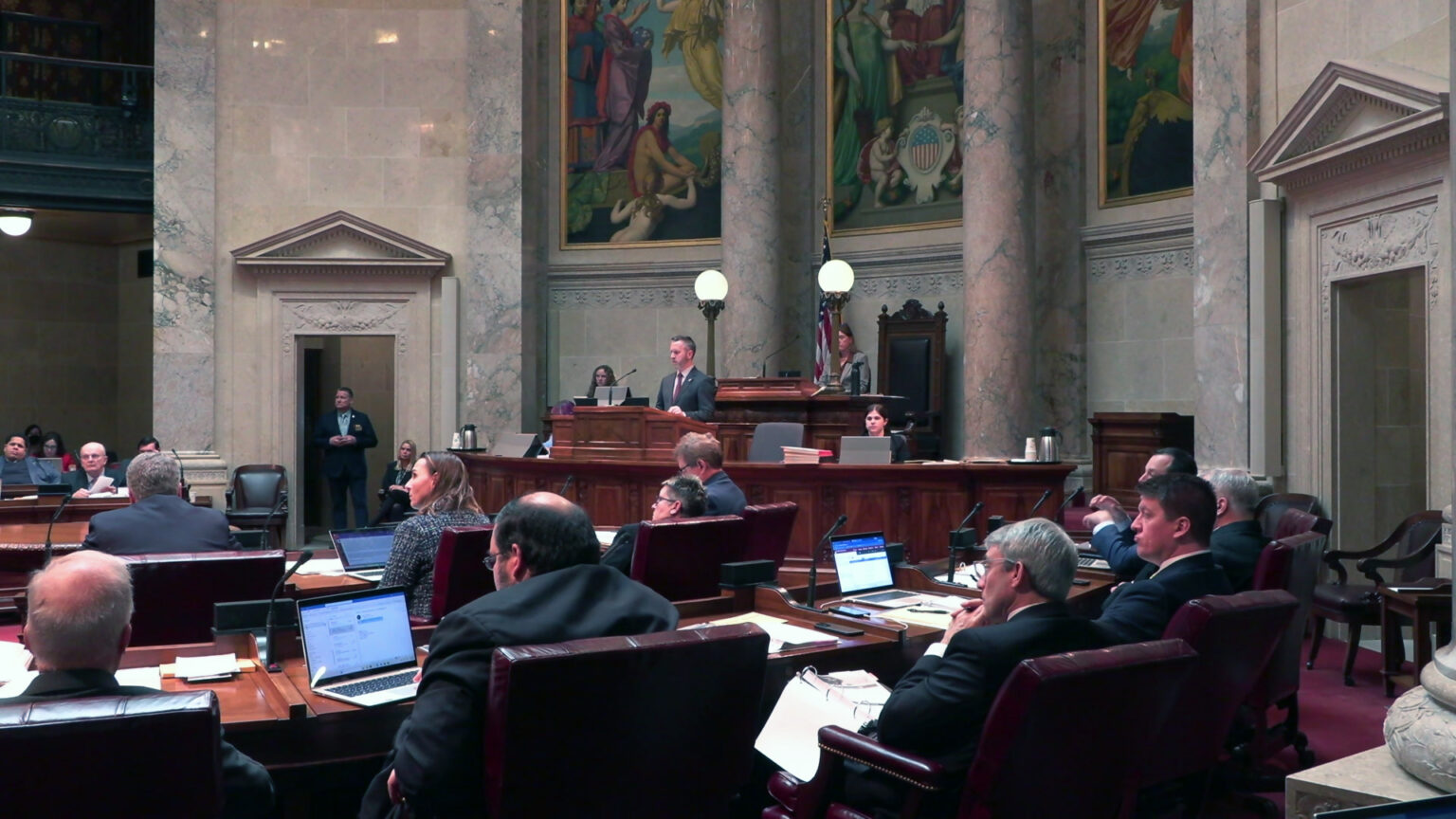 State senators sit at wood desks arranged in a semi-circle while other staff sit and stand at a wooden legislative dais, in a room with marble masonry and pillars, large paintings, and a second-story observation gallery.
