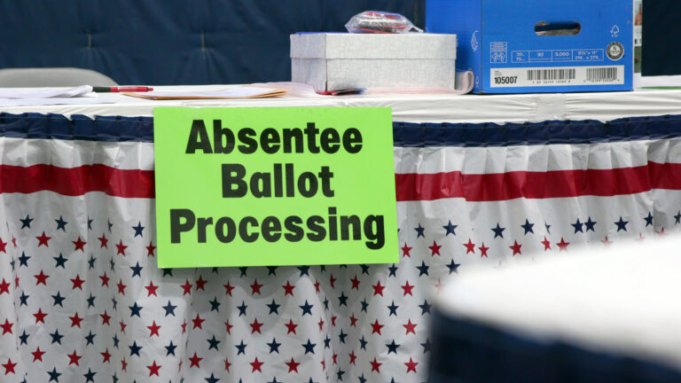 A printed sign with the words Absentee Ballot Processing is attached to the edge of a table with a red-white-and-blue stripes and stars tablecloth, with a banker's box and smaller box on its surface alongside pieces of paper and a pen. A printed sign with the words Absentee Ballot Processing is attached to the edge of a table with a red-white-and-blue stripes and stars tablecloth, with a banker's box and smaller box on its surface alongside pieces of paper and a pen.