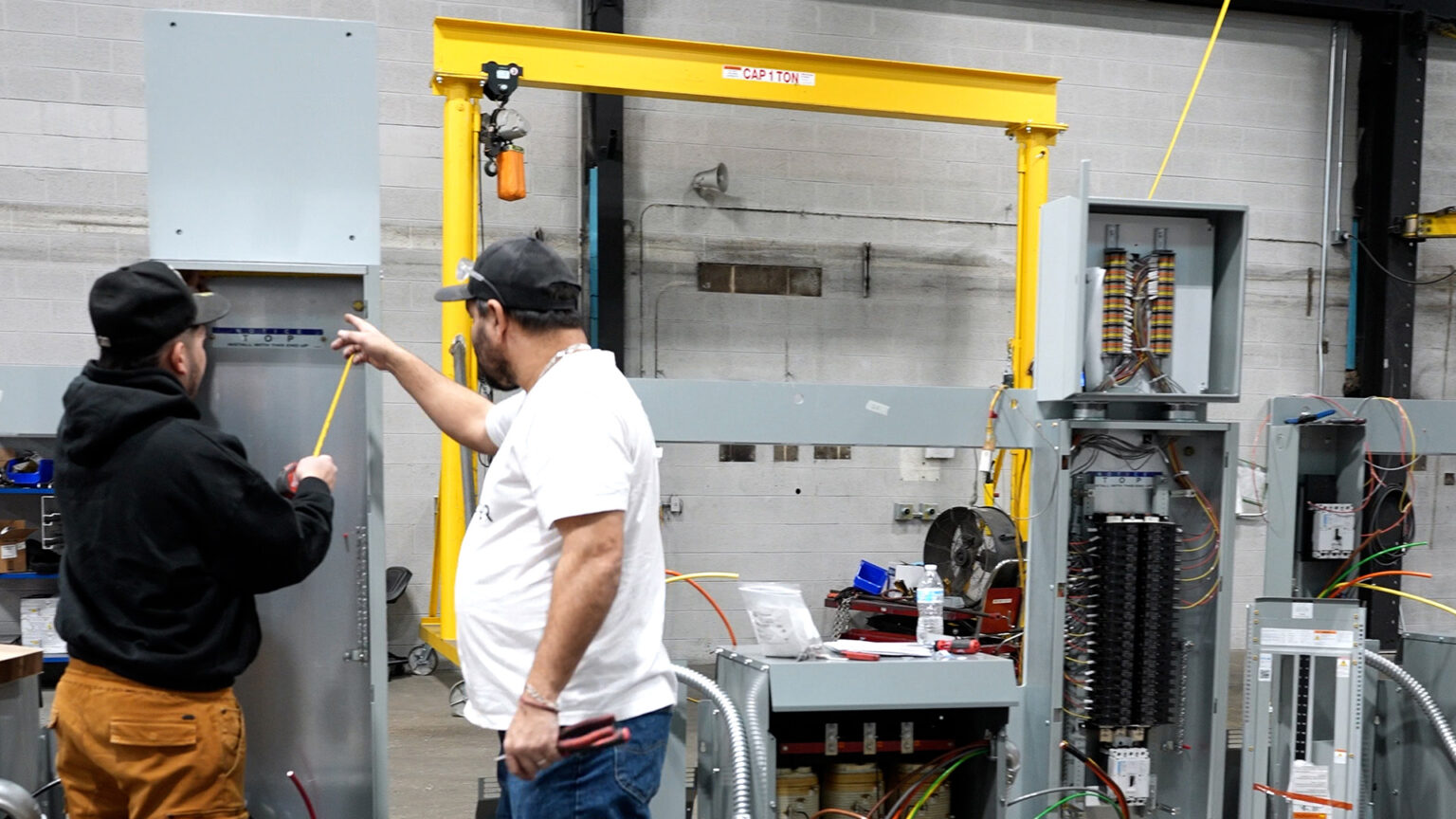One worker uses a tape measure while standing next to another worker pointing with their right index finger as both face a metal cabinet in a room with concrete block walls and multiple electrical equipment items. One worker uses a tape measure while standing next to another worker pointing with their right index finger as both face a metal cabinet in a room with concrete block walls and multiple electrical equipment items.